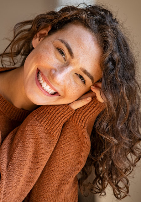 Woman with curly hair smiling at the camera while leaning her head on her hand.
