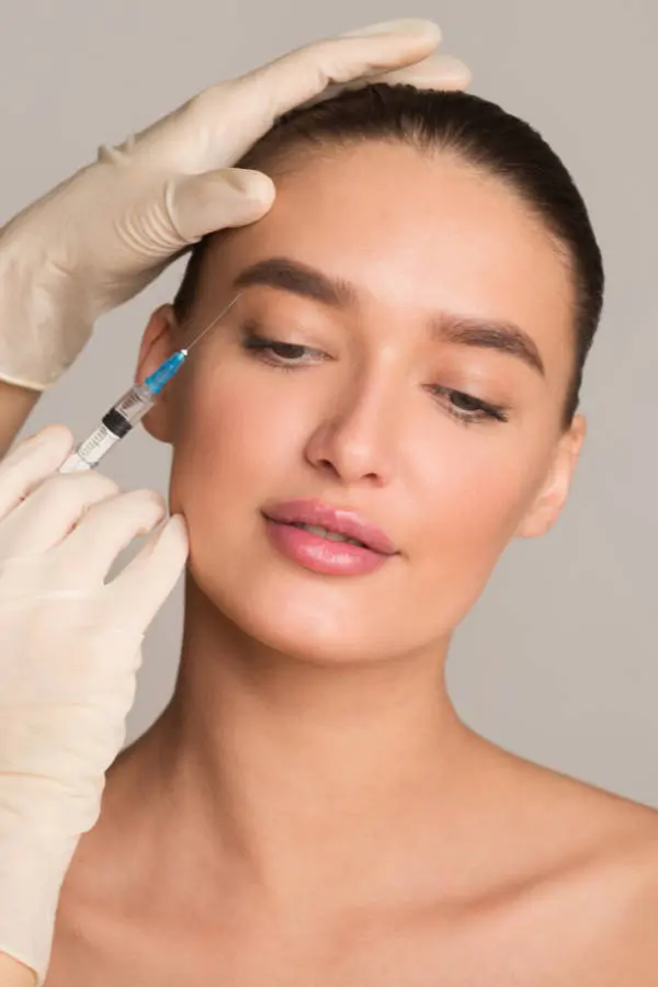 A woman receives a cosmetic injection beside her eyebrow from a gloved professional.